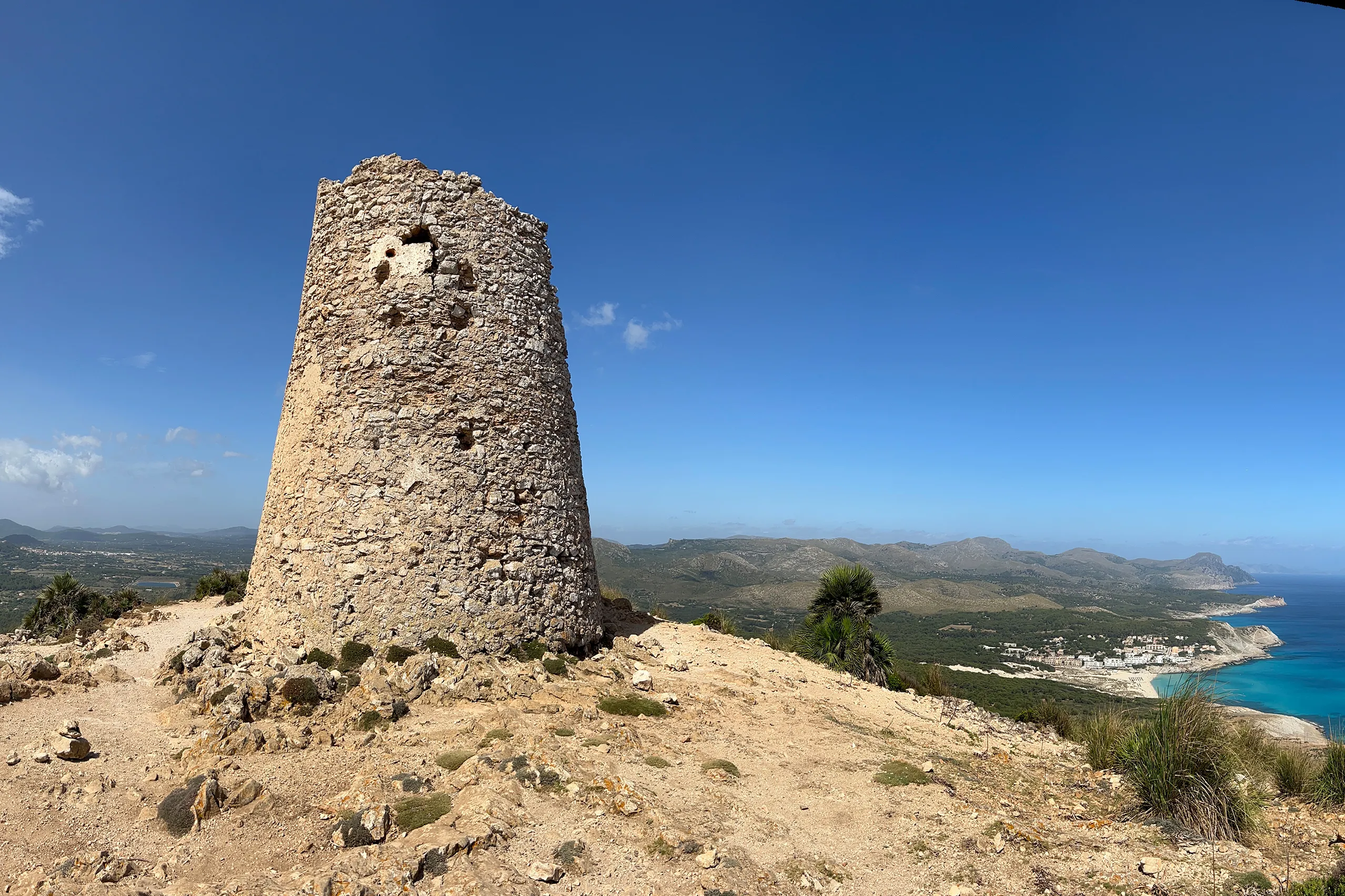 Die runde Ruine des Wachturms Torre de Son Jaumell aus grobem Bruchstein auf einem kahlen Gipfel, dahinter die bewaldeten Hügel des Hinterlands und das Meer unter blauem Himmel.
