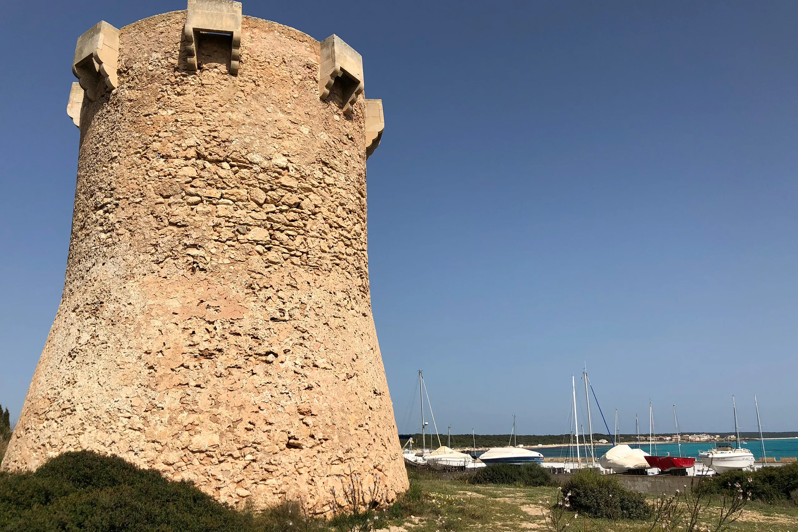 Alter runder Wachturm aus Bruchstein über dem Hafen von Sa Ràpita, dahinter Segelboote und das offene Meer unter blauem Himmel.
