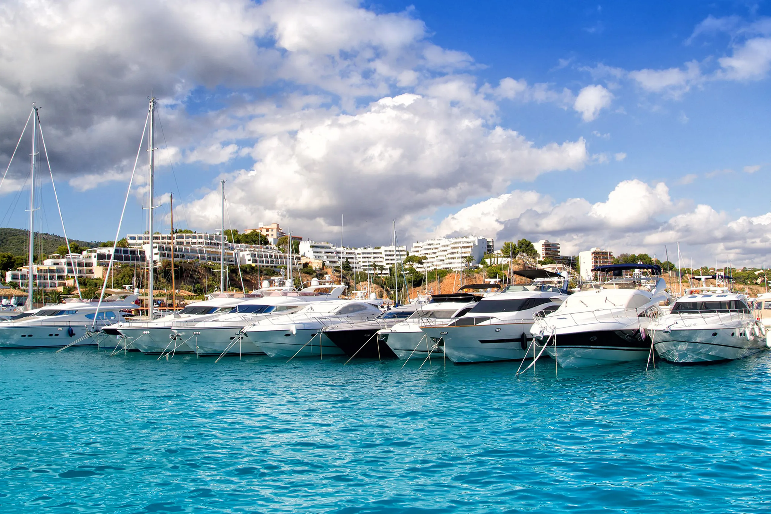 Reihe weißer Motoryachten an den Liegeplätzen von Puerto Portals, dahinter helle Apartmentbauten am bewaldeten Hang unter Wolkenhimmel.
