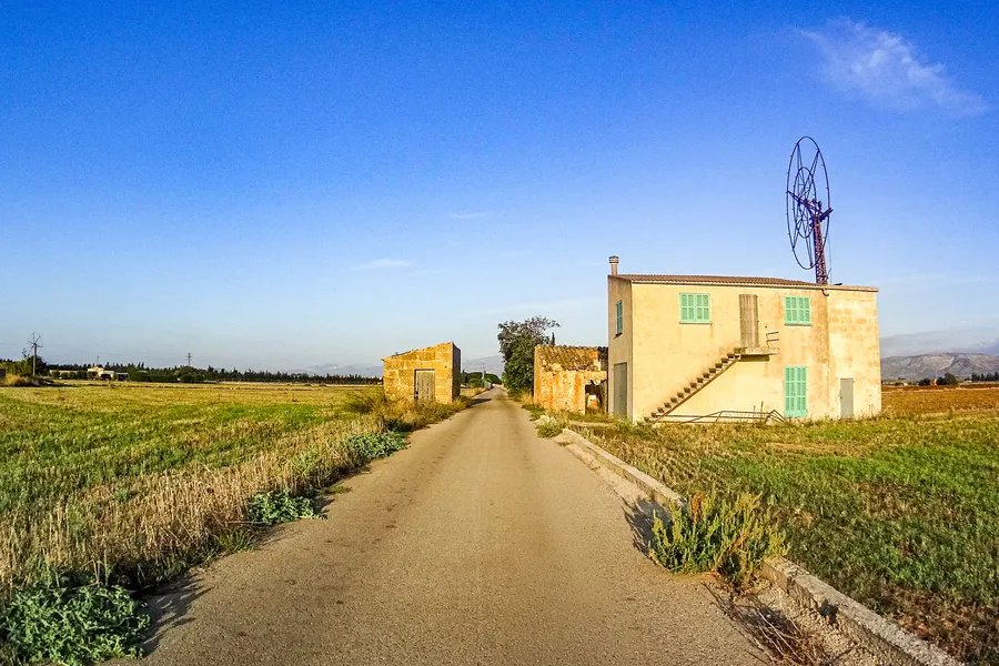 Eine leere Landstraße im Pla de Mallorca führt geradeaus durch abgeerntete Felder, rechts ein hell verputztes Landhaus mit grünen Fensterläden und einem Windrad auf dem Dach.