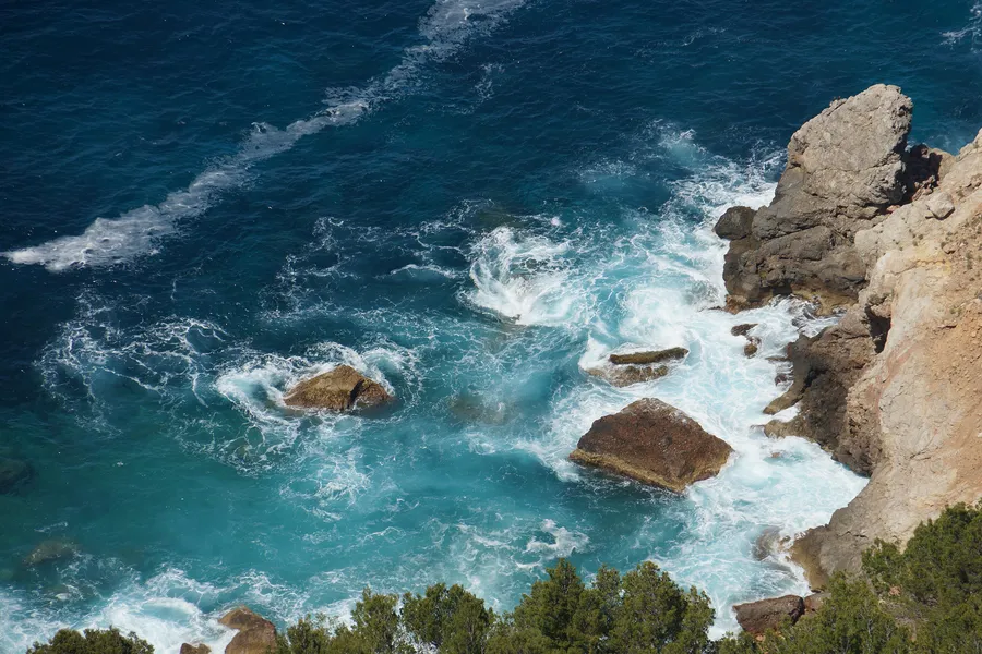 Senkrechter Blick von der Steilküste der Serra de Tramuntana hinunter auf tiefblaues Wasser, weiße Brandung und einzelne Felsblöcke im Meer.