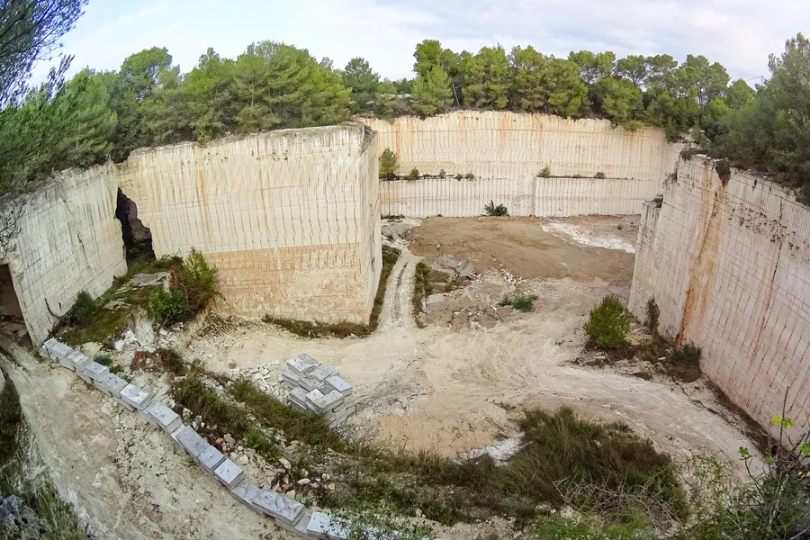 Ein aufgelassener Sandsteinbruch im Pla de Mallorca mit senkrecht geschnittenen hellen Wänden, gestapelten Steinquadern auf der Sohle und Kiefern am oberen Rand.