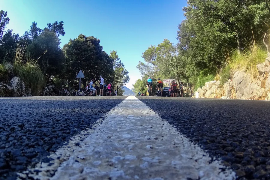 Flach über den Asphalt fotografierte Mittellinie einer Passstraße am Coll de Femenia, am Straßenrand eine Gruppe von Rennradfahrern im Schatten der Bäume.
