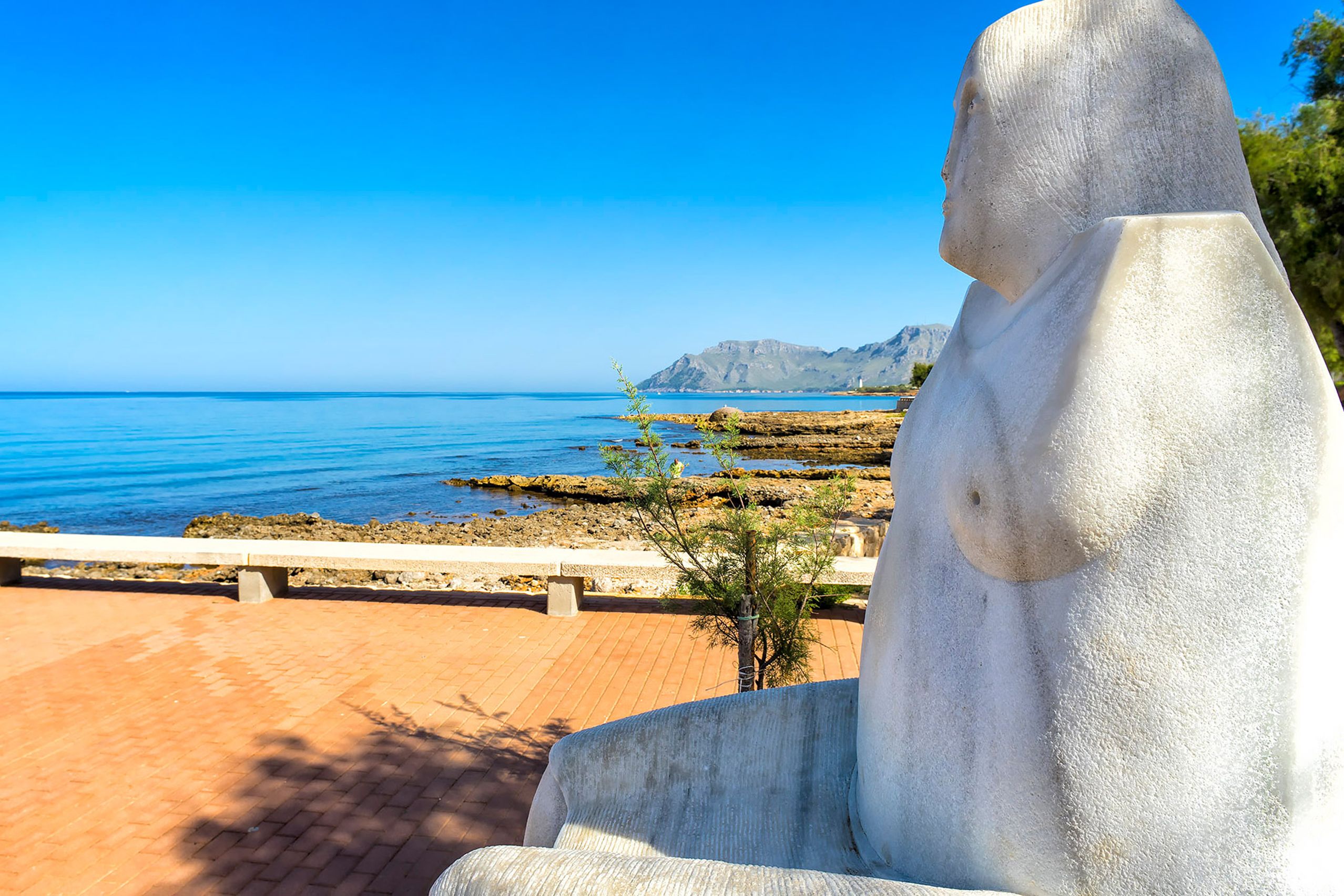 Große weiße Steinfigur einer sitzenden Frau auf der rot gepflasterten Uferpromenade, im Blick über die Felsküste hinaus auf das ruhige Meer und die Berge der Halbinsel Victòria.
