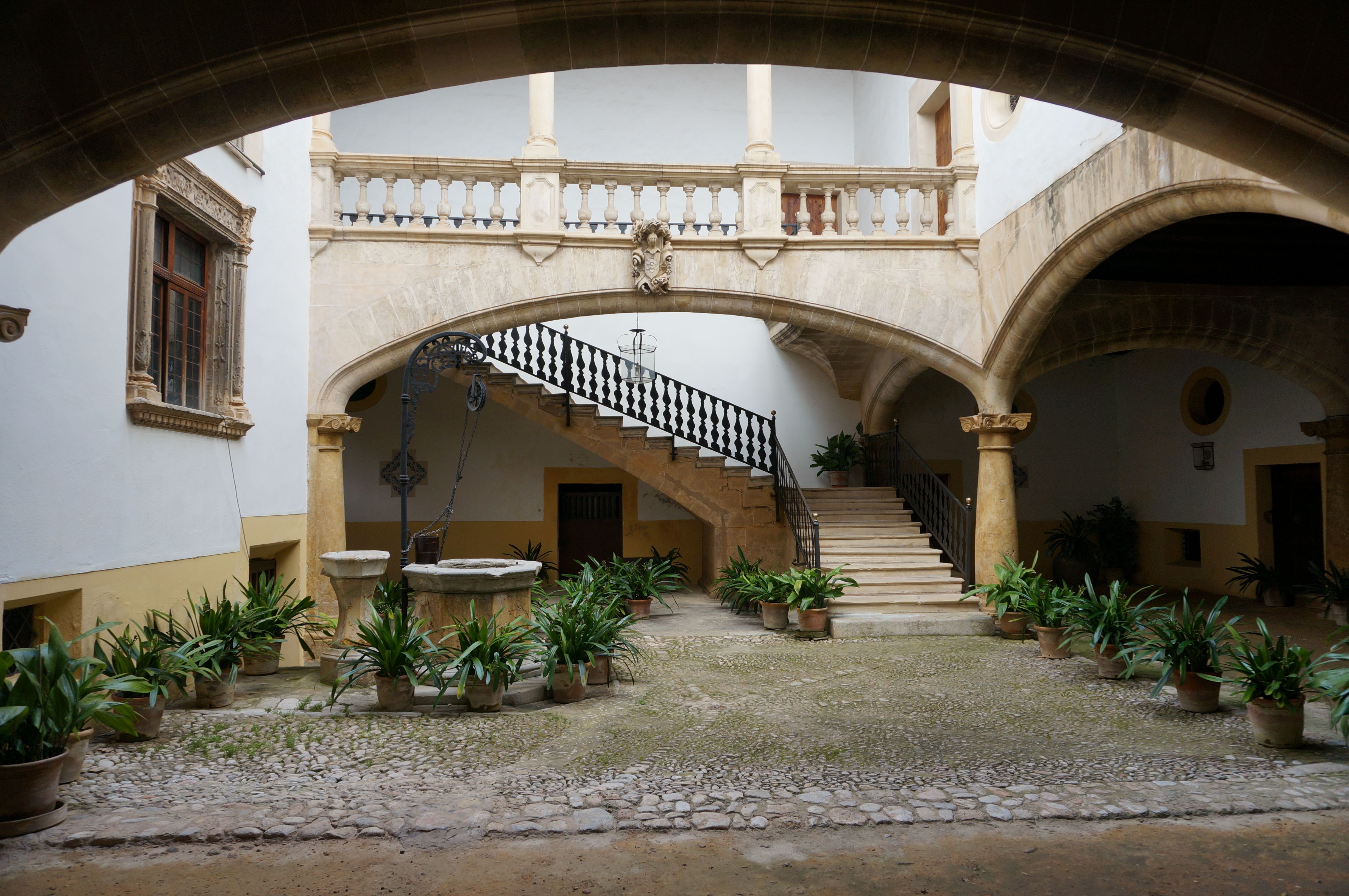 Innenhof eines Stadtpalastes in Palma mit Rundbögen, steinerner Freitreppe, schmiedeeisernem Geländer und Topfpflanzen auf Kopfsteinpflaster.
