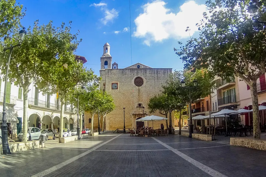 Die Plaça von Alaró mit der Pfarrkirche Sant Bartomeu, davor Platanen, Sonnenschirme der Lokale und geparkte Fahrräder.
