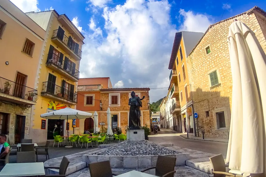 Kleiner Platz in Alaró mit Bronzedenkmal, Café-Terrasse mit geschlossenen Schirmen und Sandsteinhäusern mit grünen Fensterläden.
