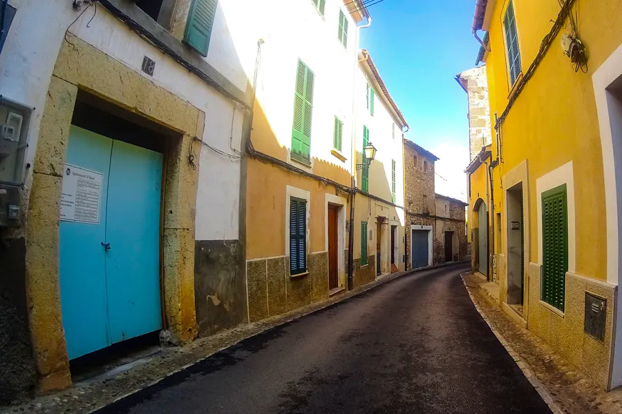 Schmale Gasse in Alaró mit hellen Häusern, türkisfarbenem Tor und grünen Fensterläden unter blauem Himmel.
