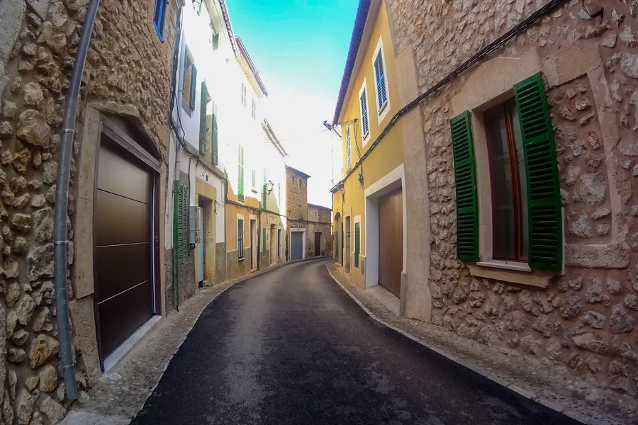 Enge, leicht ansteigende Gasse in Alaró zwischen hohen Häuserwänden mit grünen Fensterläden, am Ende Grün und Himmel.
