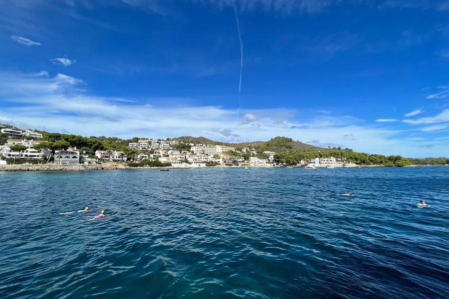 Blick über das ruhige Wasser der Bucht auf die flache, bewaldete Küste von Alcanada mit vereinzelten weißen Häusern zwischen Kiefern.