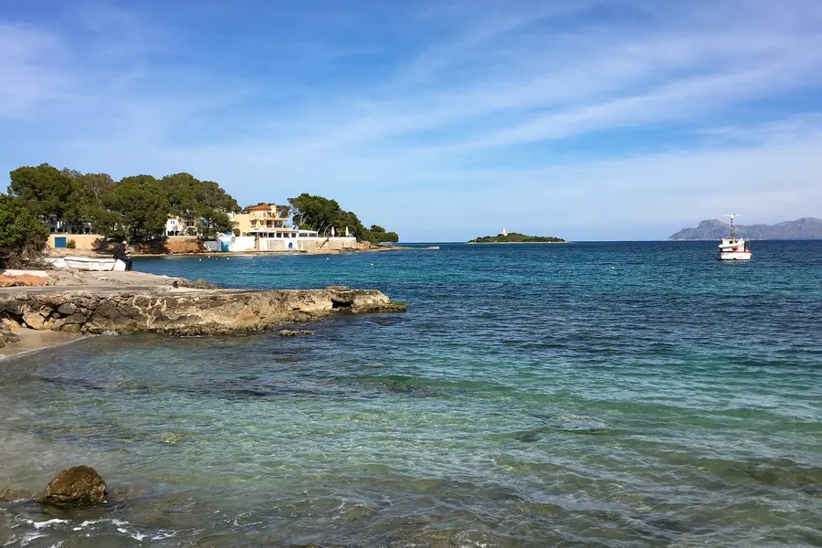 Flaches Felsufer bei Alcanada mit sehr klarem, türkisfarbenem Flachwasser, dahinter eine bewachsene Landzunge mit einzelnen Häusern und die Leuchtturminsel.