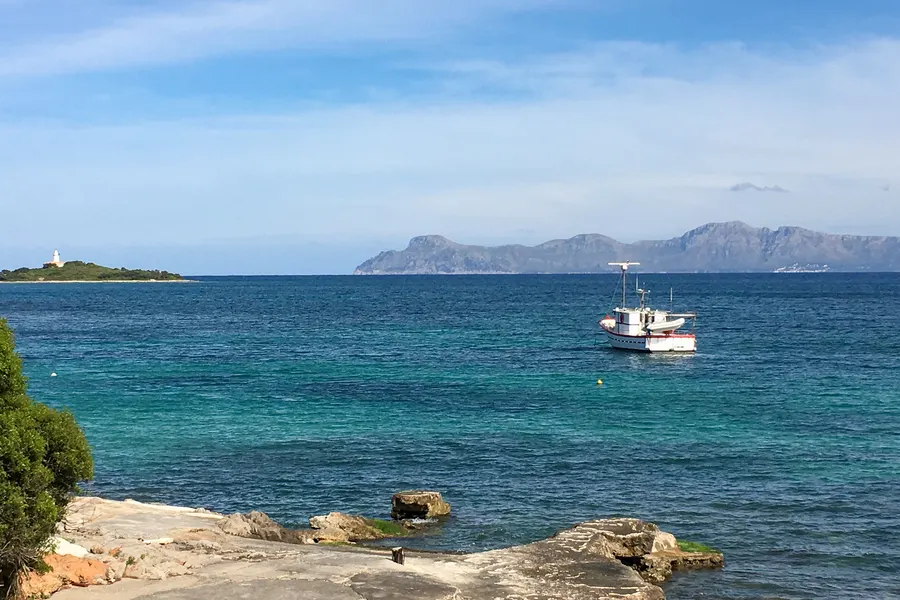 Blick über das ruhige Wasser der Bucht von Alcúdia mit einem ankernden Fischerboot, links die Leuchtturminsel, am Horizont die Berge der Halbinsel.
