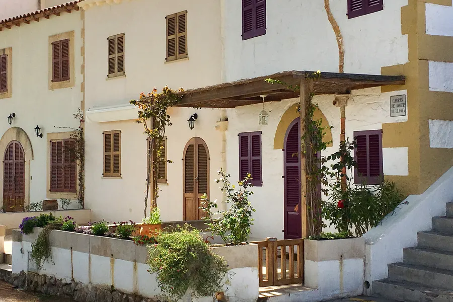 Weißes Haus mit weinroten Fensterläden und einer hölzernen Pergola über der Terrasse in Alcanada, davor bepflanzte Beete.
