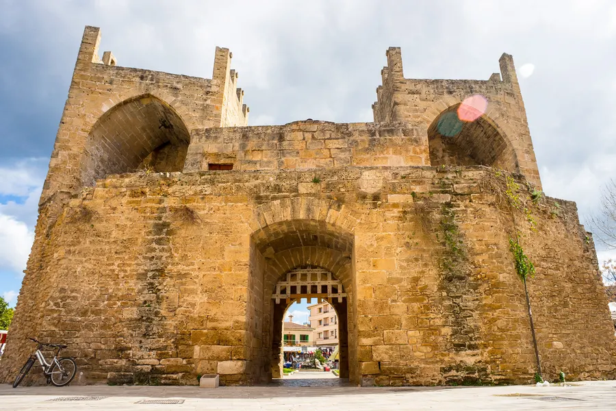Das mächtige Stadttor von Alcúdia von außen, aus grobem Sandstein gemauert, durch den Torbogen sieht man in die belebte Gasse der Altstadt.