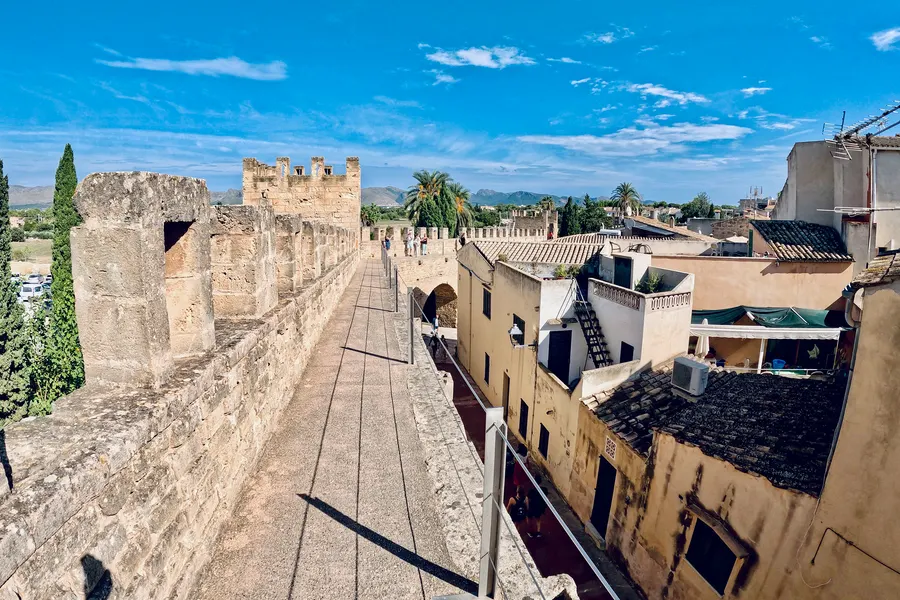 Begehbarer Wehrgang auf der Stadtmauer von Alcúdia mit Zinnen, rechts die Ziegeldächer der Altstadt, im Hintergrund die Berge der Serra de Tramuntana.