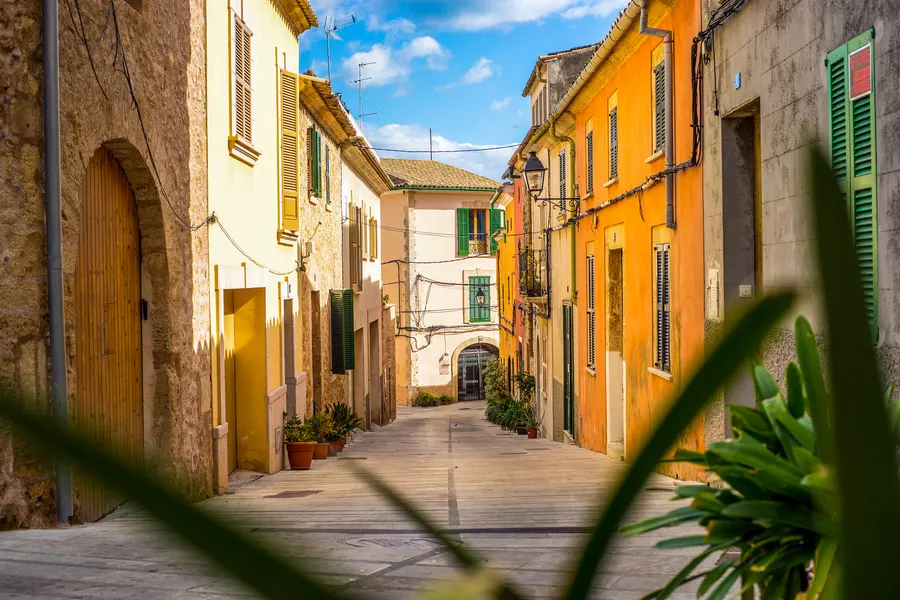 Enge Gasse in der Altstadt von Alcúdia mit ockerfarbenen und orangenen Hausfassaden, grünen Fensterläden und Topfpflanzen vor den Türen.