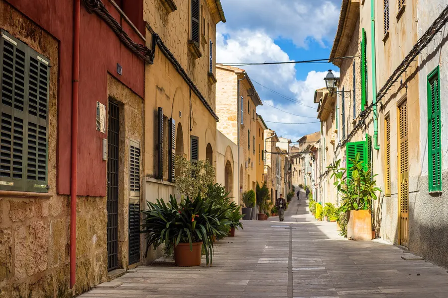 Leicht ansteigende Gasse in Alcúdia mit gepflastertem Weg, Kübelpflanzen an den Hauswänden und einzelnen Passanten in der Ferne.
