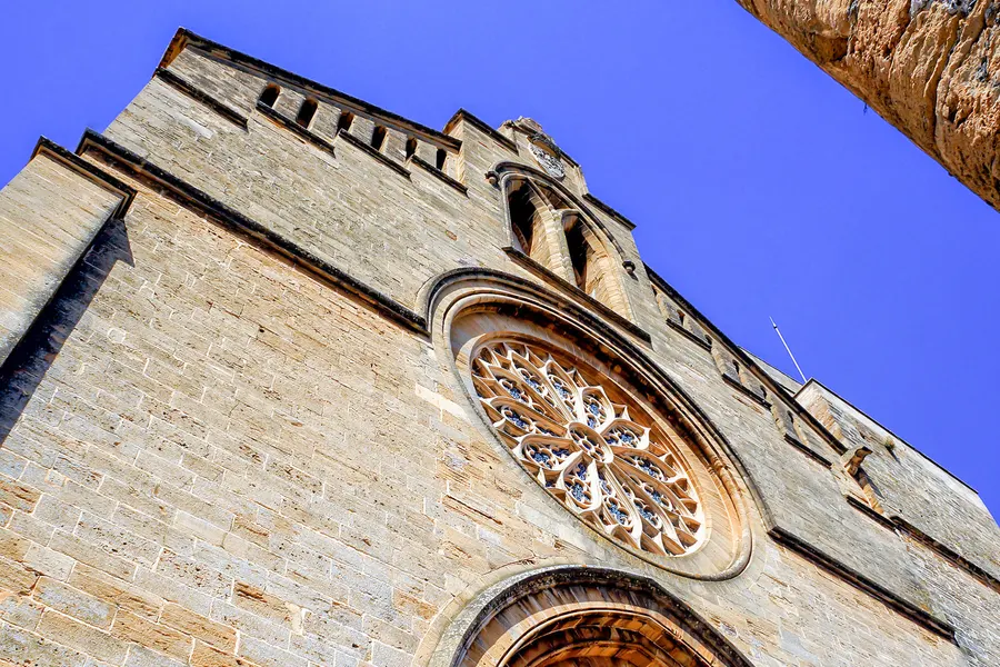 Die Fassade der Pfarrkirche Sant Jaume in Alcúdia von unten fotografiert, mit gotischem Maßwerk-Rosettenfenster über dem Portal vor tiefblauem Himmel.

