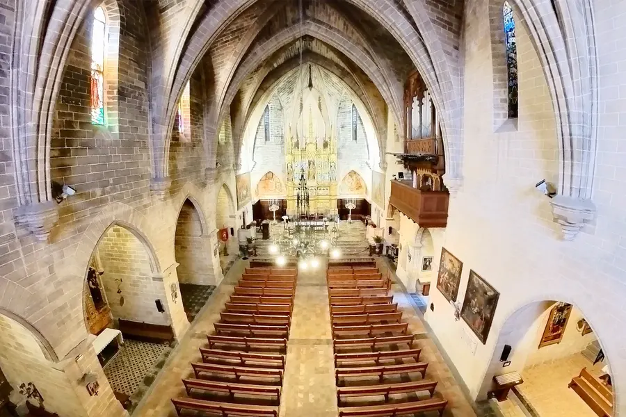 Innenraum der Kirche Sant Jaume in Alcúdia mit hohen Spitzbögen, hellem Gewölbe, Holzbänken im Mittelgang und dem beleuchteten Altar im Chor.
