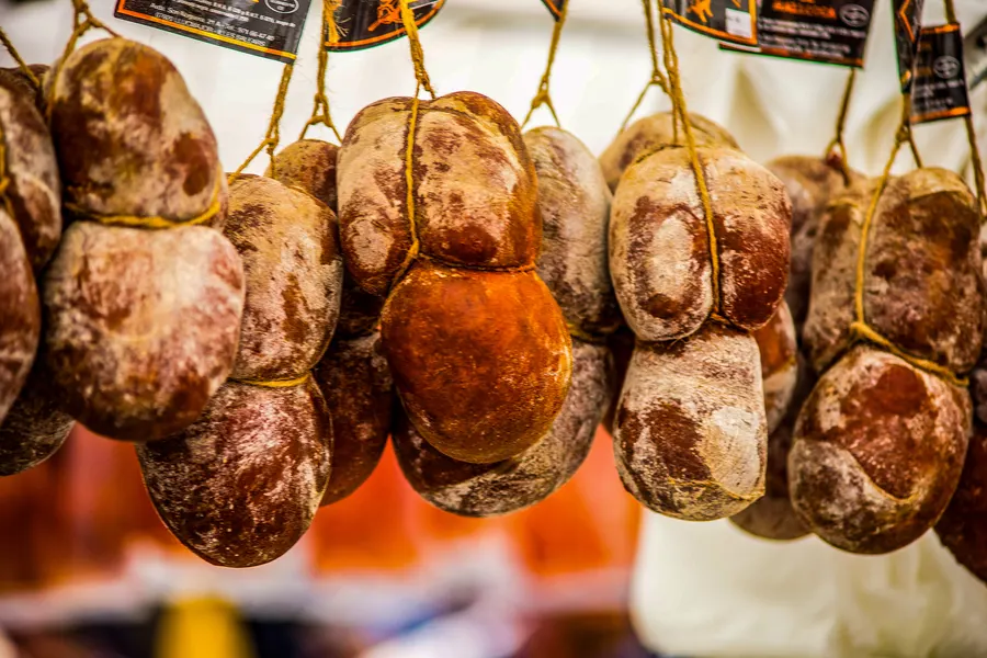 An Schnüren hängende Sobrassada-Würste in Reihe an einem Marktstand in Alcúdia, im Hintergrund unscharf die Marktplane.
