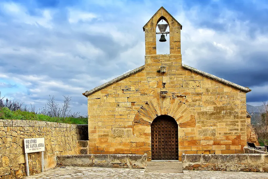 Die kleine gotische Kapelle Oratori de Santa Anna bei Alcúdia aus hellem Sandstein, mit Rundbogenportal und Glockengiebel über dem Dachfirst.
