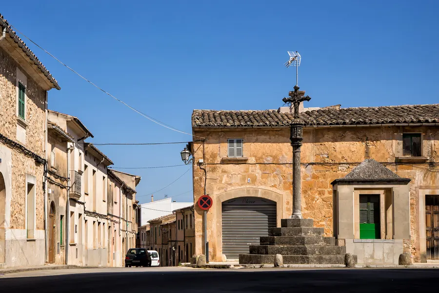 Das steinerne Renaissance-Wegkreuz Creu des Colomer auf gestuftem Sockel an der Durchfahrtsstraße von Algaida, dahinter niedrige Häuser und eine Kapelle.