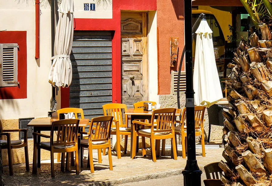 Caféterrasse in Andratx mit hellen Holzstühlen und geschlossenen weißen Schirmen vor einer rot gestrichenen Fassade, daneben ein Palmstamm.
