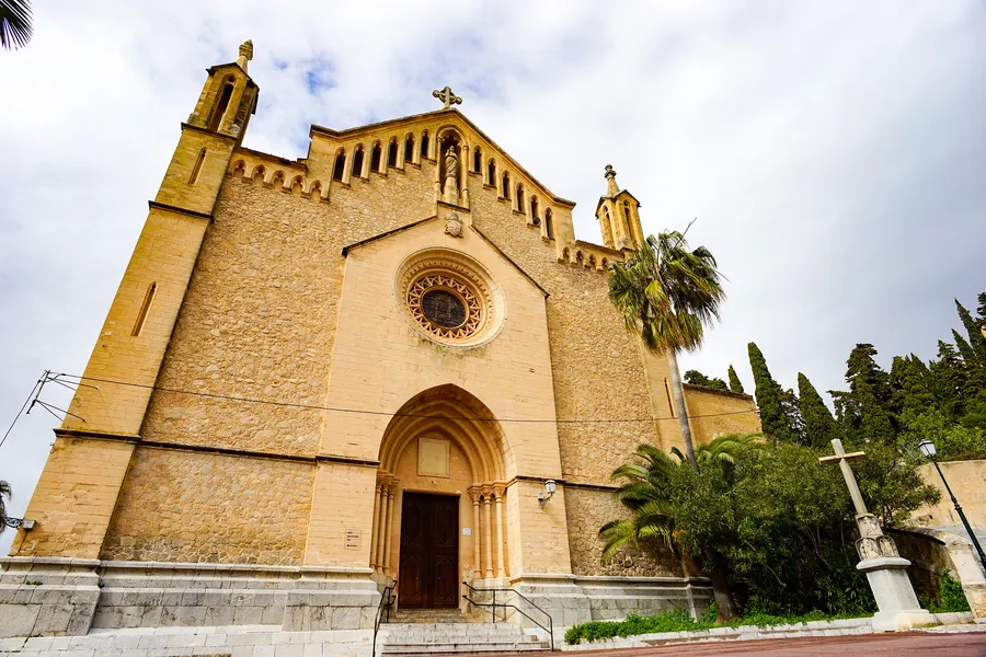 Die Pfarrkirche Transfiguració del Senyor in Artà von vorn: hoher Sandsteingiebel mit Rosettenfenster über dem Portal, links und rechts Palmen und Zypressen.