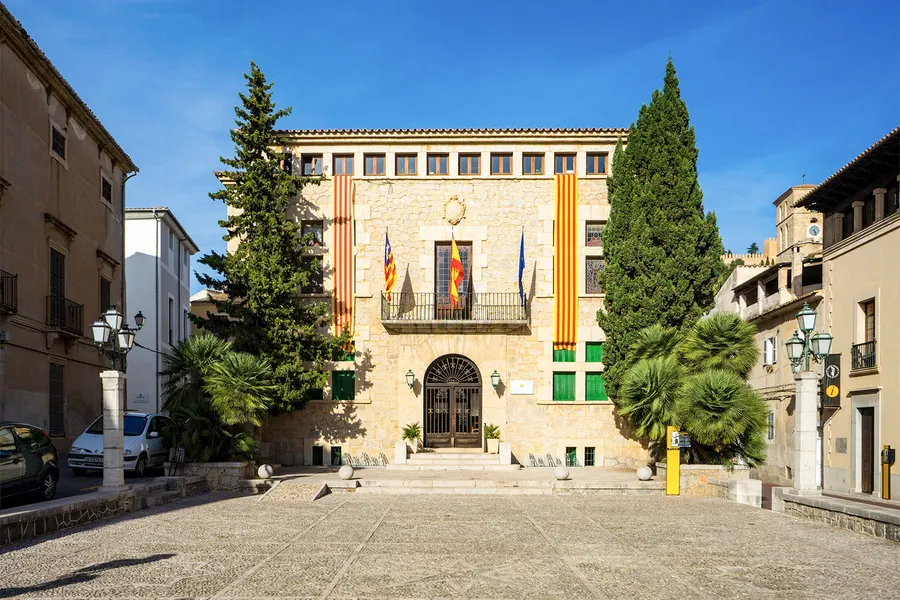 Das Rathaus von Artà an einem gepflasterten Platz, ein heller Sandsteinbau mit Fahnen über dem Portal, davor Zypressen und Straßenlaternen.
