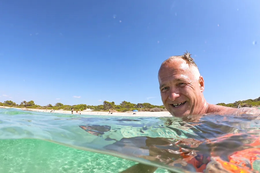 Friedhelm Fischer schwimmt im klaren, flachen Wasser vor der Platja des Carbó, im Hintergrund der weiße Sandstrand mit Kiefern.