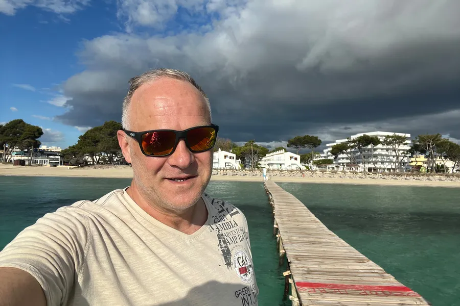 Friedhelm Fischer mit Sonnenbrille auf einem Holzsteg an der Platja de Muro, dahinter der Strand mit Liegen und Hotels unter aufziehenden Wolken.