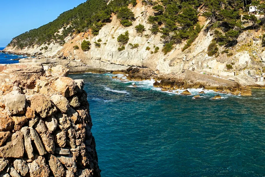 Die Bucht am Fuß von Banyalbufar mit hoher heller Felswand, türkisfarbenem Wasser und einem schmalen Kiesstreifen.