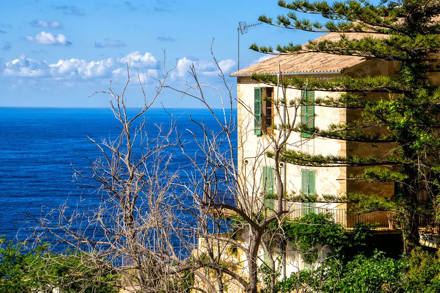 Haus mit grünen Fensterläden über der Steilküste von Banyalbufar, davor ein kahler Baum und tiefblaues Meer.
