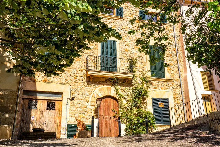 Ockerfarbenes Natursteinhaus in Banyalbufar mit grünen Fensterläden, schmiedeeisernem Balkon und Weinlaub über der Fassade.
