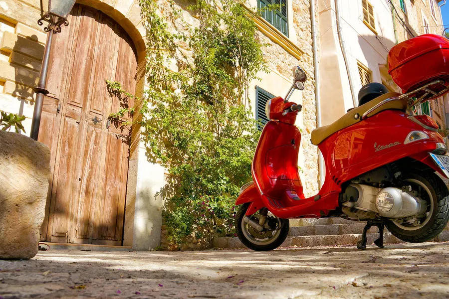 Rote Vespa vor hölzernen Toren an einer ansteigenden Gasse in Banyalbufar, daneben Grün und Natursteinmauern.
