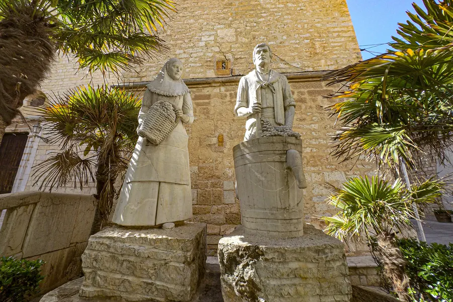 Zwei helle Steinfiguren vor der Kirche von Binissalem: eine Frau mit einem Korb und ein Mann, der auf einem Fass steht, umgeben von Palmen.
