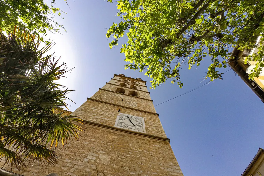 Der Glockenturm von Binissalem mit heller Zifferblattuhr, steil von unten durch Palmwedel und Platanenblätter fotografiert.

