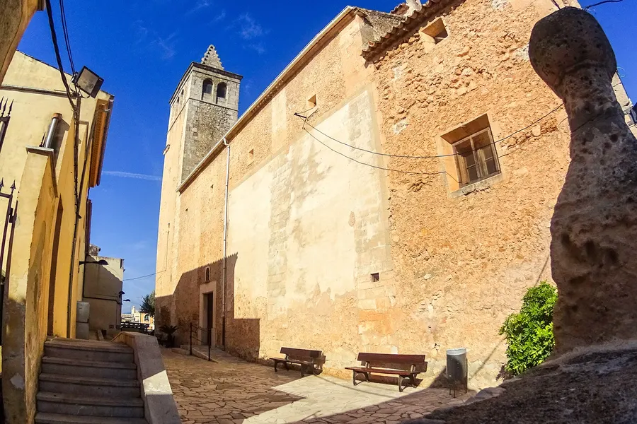 Seitenansicht der Kirche Sant Pere in Búger mit verwitterter Natursteinfassade und Strebepfeilern, daneben die Gasse mit Wohnhäusern.