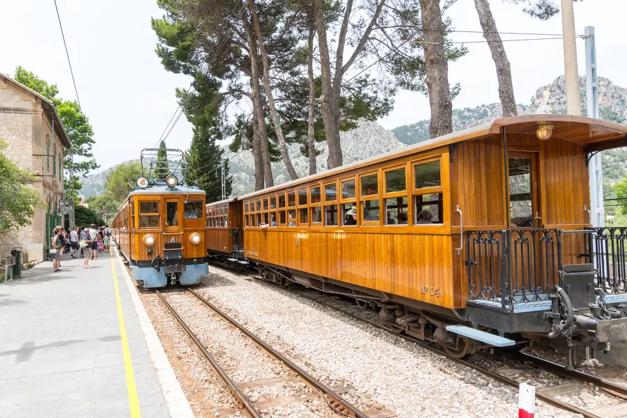 Triebwagen und angehängte Holzwagen am Bahnsteig von Bunyola, im Hintergrund wartende Reisende und die Bergflanke.
