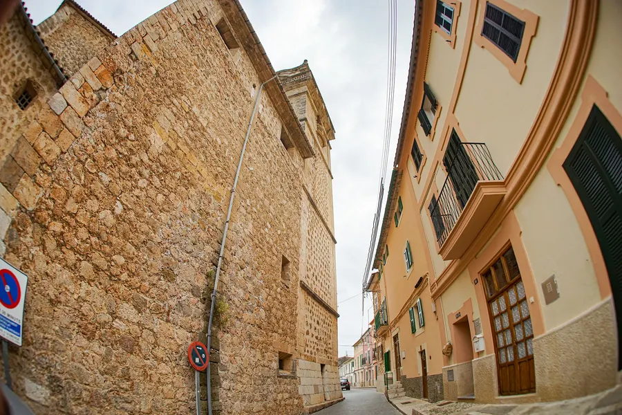 Enge Gasse in Bunyola, von unten fotografiert, zwischen einer hohen Bruchsteinmauer und Häusern mit Balkonen.
