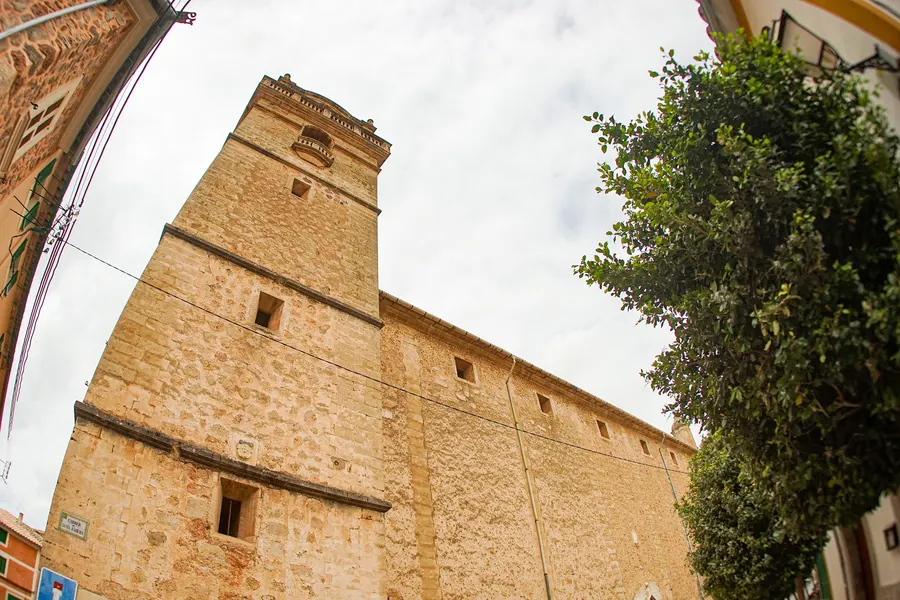 Die hohe, fast fensterlose Sandsteinwand der Kirche von Bunyola, steil von unten fotografiert, daneben ein Baum.
