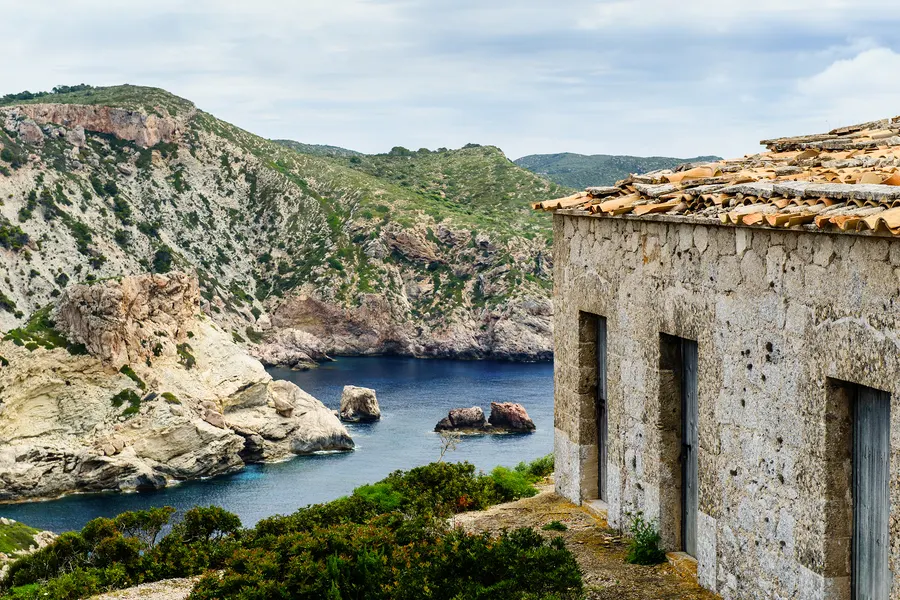 Verlassenes Steinhaus mit leeren Fensteröffnungen an einer Bucht auf Cabrera, dahinter Felsküste und Meer.