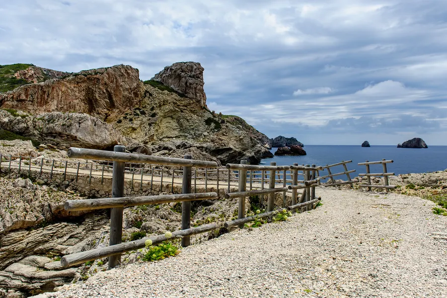 Geschotterter Wanderweg mit Holzgeländer entlang der Felsküste von Cabrera, dahinter Meer und vorgelagerte Inselchen.
