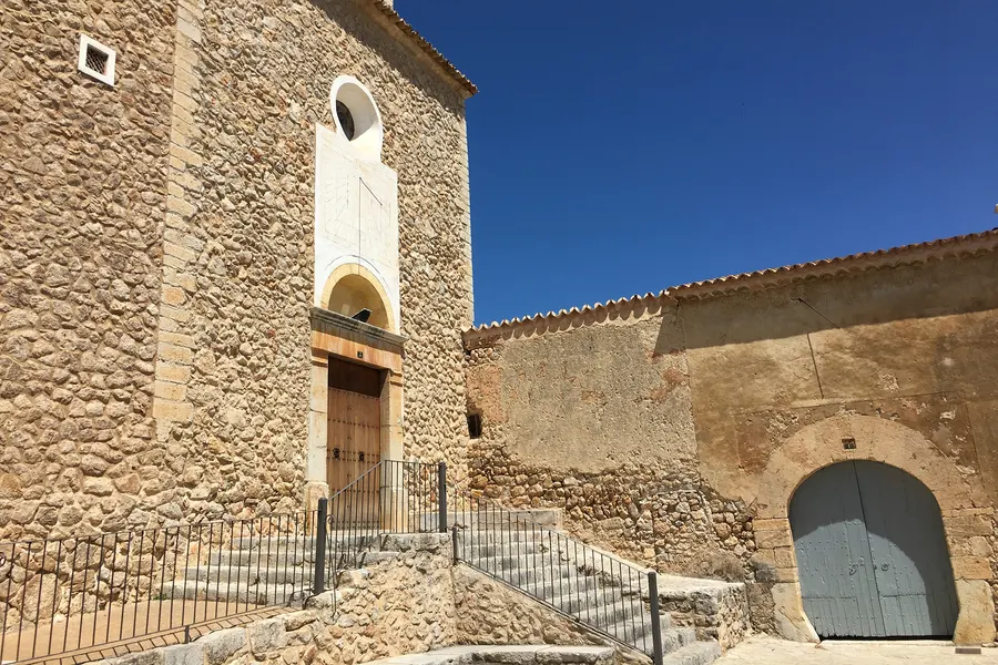 Schlichte Kirchenfassade aus Bruchstein in Caimari mit Rundfenster, Glockengiebel und einer Freitreppe mit Geländer davor.