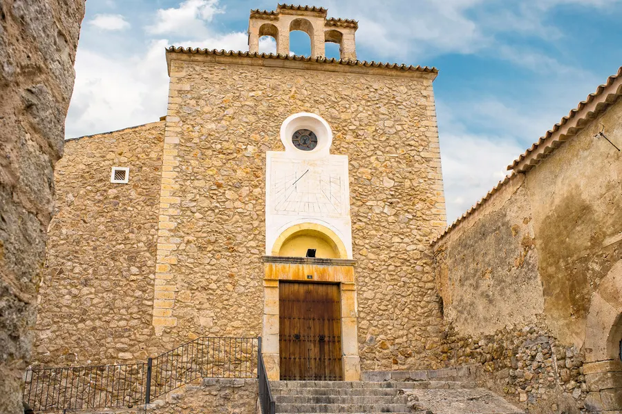 Seitenansicht der Kirche von Caimari mit hoher Bruchsteinmauer, Glockengiebel, Sonnenuhr und angebautem Wirtschaftsgebäude.