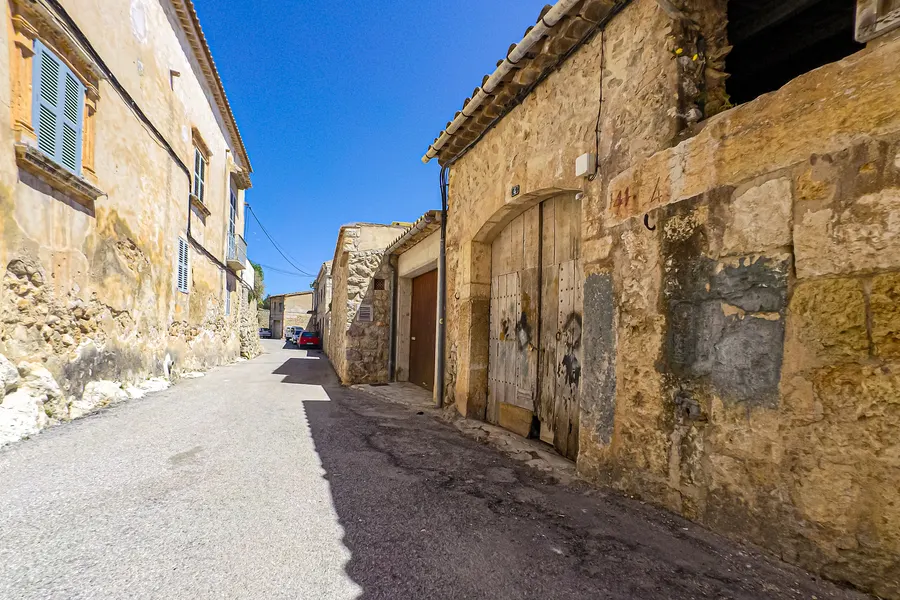 Straße in Caimari mit alten Wirtschaftsgebäuden, breiten Holztoren und einer verwitterten Steinfassade.

