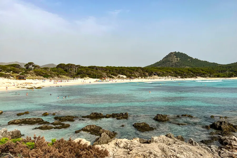 Sehr klares türkisfarbenes Flachwasser mit dunklen Felsbänken, dahinter der helle Strandbogen und ein bewaldeter Hügel.