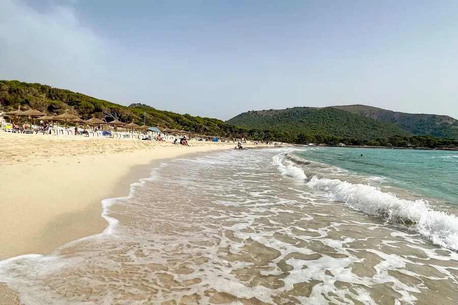Auslaufende Wellen auf dem hellen Sand der Cala Agulla, dahinter der besetzte Strand mit Liegen und die bewaldete Landzunge.