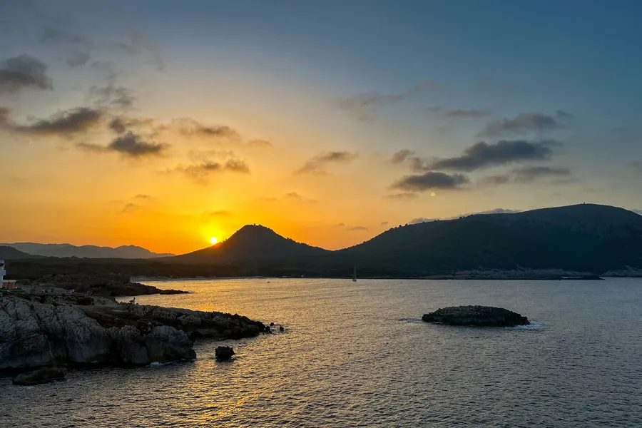 Sonnenuntergang hinter den Hügeln über der Bucht bei Cala Agulla, orangefarbener Himmel, dunkle Silhouetten der Landzungen und ruhiges Wasser.
