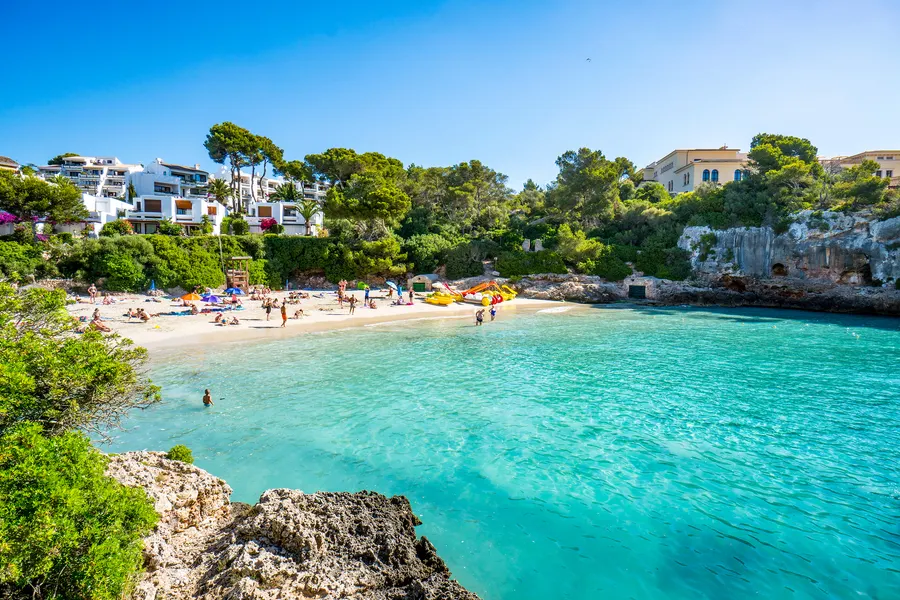 Die Cala Gran mit hellem Sandstrand, Badegästen im türkisfarbenen Flachwasser und Kiefern auf den Felsrändern.